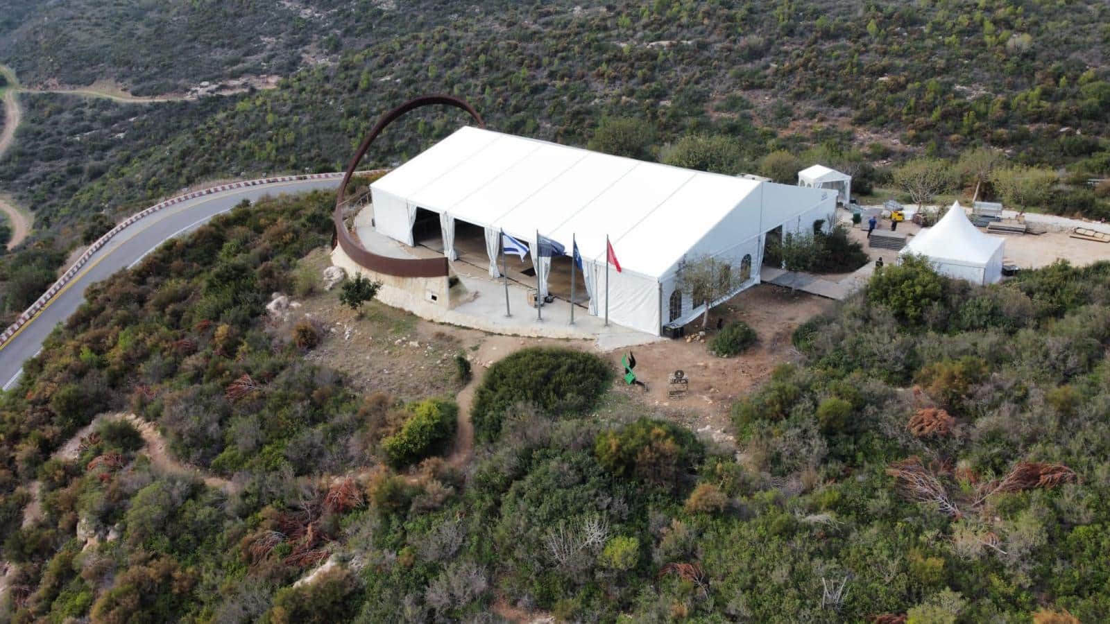 מבנה לבן גדול דמוי אוהל הממוקם על ראש הר במרחב טבעי מוקף צמחייה, סמוך לעיקול כביש הררי, ולצידו מתנוססים דגלים ופסל מתכתי קשתי בולט.