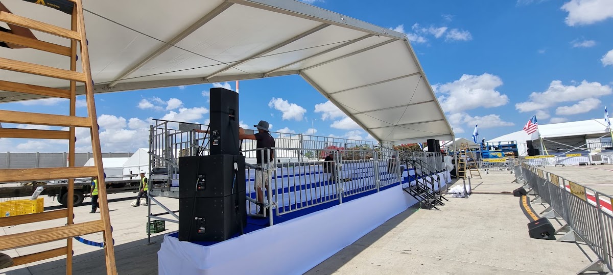הקמת אוהל אירועים גדול עם במות וציוד סאונד לאירוע חיצוני תחת שמיים בהירים.